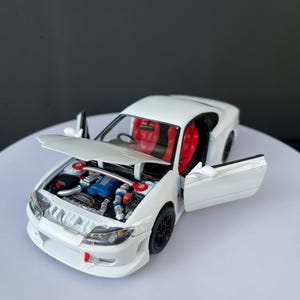 Op de afbeelding: Een wit gegoten modelauto met een geopende motorkap en deuren. Het interieur is voorzien van felrode kuipstoelen. De motorruimte is gedetailleerd met een blauw motorblok, zilverkleurige leidingen en rode veerpootbruggen. Zwarte wielen.