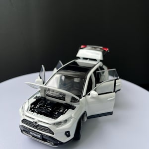 May include: A white die-cast model SUV with a black roof, open doors, and hood. The model car has the word "RAV4" on the front. The car's doors, hood, and rear hatch are open, revealing the interior and engine details.