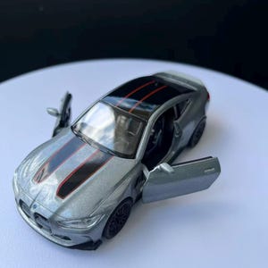 May include: A silver-gray model car with black and red stripes on the hood and roof. The doors are open, revealing the interior. The car has black wheels and a black roof.