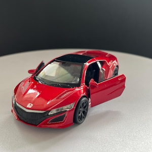 May include: A red die-cast model car featuring a black roof, black wheels, and a Honda emblem on the front hood. The vehicle has two open doors, clear windshields, and detailed headlights, capturing the sleek design of a sports car in miniature.