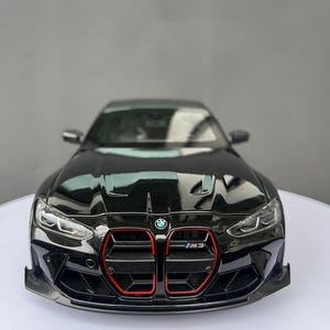 Puede incluir: Un coche a escala BMW M3 de color negro con una prominente parrilla vertical con bordes rojos, faros LED afilados y un pequeño emblema de BMW en el capó. El parachoques delantero incluye grandes tomas de aire y un divisor frontal negro.