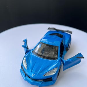 May include: A blue die-cast model car with open doors. The toy car has a sleek design, visible headlights, and a rear spoiler. The car is approximately 15 cm long.