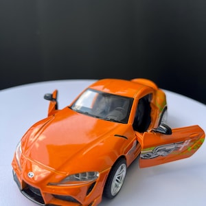 以下が含まれることがあります： ドアが開いたオレンジ色のダイキャスト製モデルカー。おもちゃの車は、詳細なインテリア、シルバーのホイール、前面のトヨタエンブレムが特徴です。車にはドアにグラフィックデザインがあります。車は白い表面にあります。