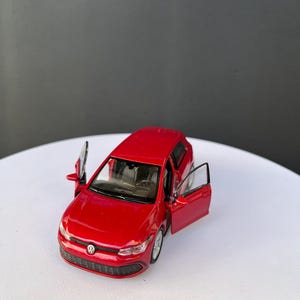 May include: A bright red die-cast Volkswagen Golf model car featuring a detailed exterior with a front grille emblem, silver wheels, and two open front doors that reveal a dark interior cabin with a steering wheel and dashboard components.