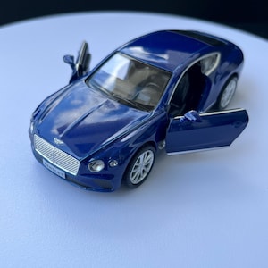 Może przedstawiać: Szczegółowy model niebieskiego Bentley Coupe z otwartymi drzwiami. Miniaturowy samochód ma srebrne koła, czarne wnętrze i logo Bentley z przodu. Przedmiot kolekcjonerski.