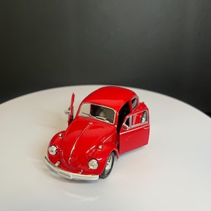 May include: A bright red die-cast model of a VW Beetle car featuring a glossy finish, silver-toned bumpers, and round headlights. The driver and passenger side doors are open, revealing the interior, and a small license plate reads VW Beetle.
