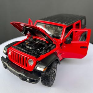 Könnte beinhalten: Ein rotes Jeep Rubicon Modellauto mit schwarzem Dach, schwarzen Kotflügeln und schwarzen Rädern. Die Motorhaube ist geöffnet und gibt den Blick auf den Motorraum frei, die Fahrertür ist offen und zeigt die Details des Miniatur-SUVs.