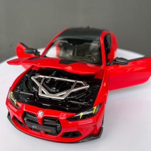 May include: A red BMW M4 scale model car with a black roof, open doors, and an open hood revealing the engine bay. The front grille features the BMW logo and an M4 emblem. The interior is visible through the open doors, and the car has detailed headlights with yellow accents.