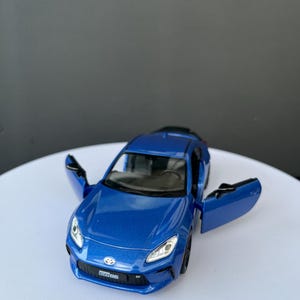 May include: A blue Toyota GR86 die-cast model car with both doors open. The vehicle features a front grille with the GR86 logo, clear headlights, and a metallic blue paint finish. The interior is visible through the windshield and open doors.