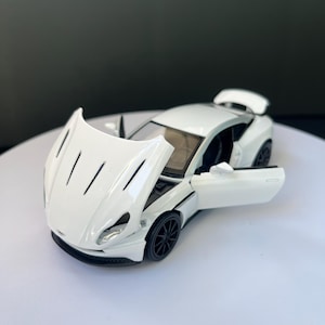 May include: A white die-cast model sports car featuring a sleek aerodynamic body, black multi-spoke wheels, and a rear spoiler. The front hood is open, revealing the engine bay, and the driver-side door is open, showing the interior cabin.