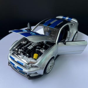 May include: Silver model car with blue racing stripes on the hood and roof. The hood and doors are open, revealing the engine and interior. The car has a yellow air filter and white wheels.