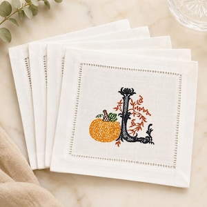May include: A stack of white square linen napkins featuring a hemstitched border. Each napkin displays an embroidered design of a bright orange pumpkin next to a large, ornate black letter L, accented with delicate orange and brown foliage.