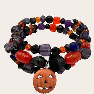 May include: A multi-strand wrap bracelet featuring a variety of black, orange, and purple beads in different shapes and textures. The bracelet includes a prominent orange circular charm with a carved jack-o'-lantern face hanging from the center.