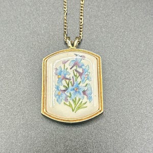 May include: A gold-tone necklace featuring a rectangular pendant with a cream-colored center. The pendant displays a painted illustration of blue flowers with green stems and leaves, enclosed within a textured gold-tone metal frame.