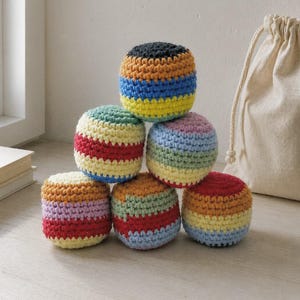 Puede incluir: Seis bolas de ganchillo a rayas multicolores, dispuestas en forma de pirámide. Las bolas presentan una variedad de colores, incluyendo rojo, amarillo, azul, verde y morado. Un saco beige con cordón se encuentra en el fondo.