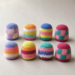 May include: Eight colorful, handmade crochet egg-shaped objects. Each egg features a unique combination of colors, including blue, pink, yellow, orange, and green. Some have striped patterns, while others have a checkered design. The eggs are arranged in two rows.