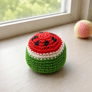 May include: A handmade crochet ball designed to resemble a watermelon. The ball features a red top with black seed details, a white stripe, and a green base. A smaller ball with pink and yellow stripes is also visible.
