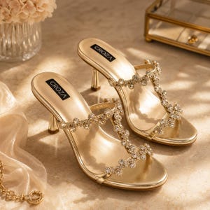 Könnte beinhalten: Ein Paar goldfarbene Sandalen mit Absatz und T-Riemen-Design, das mit klaren, funkelnden Kristallen verziert ist. Das Fußbett ist in Metallic-Gold gehalten und trägt den Markennamen CROZA in schwarzer Schrift im Fersenbereich. Der Absatz ist schmal.