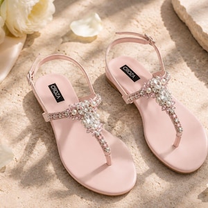 May include: A pair of light pink flat sandals featuring a T-strap design adorned with clear crystals and white faux pearls. The sandals have an adjustable ankle strap with a buckle and a black label on the insole with the brand name CROZA printed.