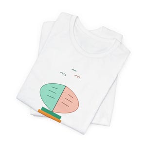 May include: A white t-shirt featuring a minimalist graphic of a split oval shape in mint green and soft peach, resting on a stacked green and orange base, with three small, simple line-drawn birds flying above the central design element.