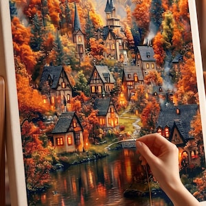 May include: A diamond painting kit on a wooden easel featuring a detailed autumn landscape with a village of houses, a winding river, and vibrant orange and yellow trees. A needle is held near the canvas, which is filled with colourful gems.