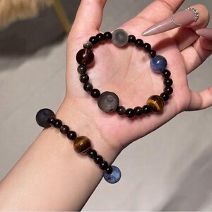 May include: Two beaded bracelets featuring a series of small black beads interspersed with larger, distinct spherical stones in various colors including brown, blue, grey, and dark red. The bracelets are constructed with elastic cord.