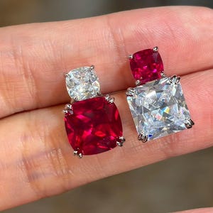 May include: Two earrings featuring a cushion-cut stone set above a larger square-cut stone. One earring has a clear cushion-cut stone atop a deep red square-cut stone, while the other has a red cushion-cut stone atop a clear square-cut stone.