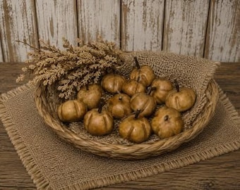 May include: A round woven basket filled with small, smooth, tan-colored decorative pumpkins. The basket is lined with a piece of brown burlap fabric and includes a cluster of dried, light brown foliage tucked into the side of the arrangement.