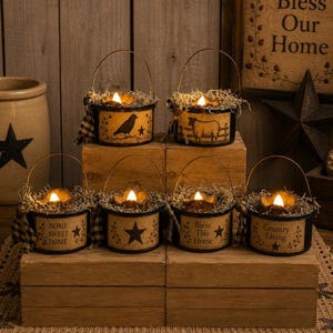 May include: Six small, round, black-rimmed metal buckets with wire handles, each containing a battery-operated candle and dried moss. The buckets feature cream-colored labels with black graphics of birds, sheep, stars, and text including "Home Sweet Home," "Bless This Home," and "Country Living."
