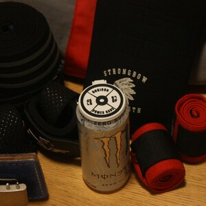 May include: An assortment of fitness equipment, including black and red wrist wraps, a weightlifting belt, and a rolled-up black band. A silver Monster Energy drink can is also present, along with a black bag with the text "STRONGBOW".