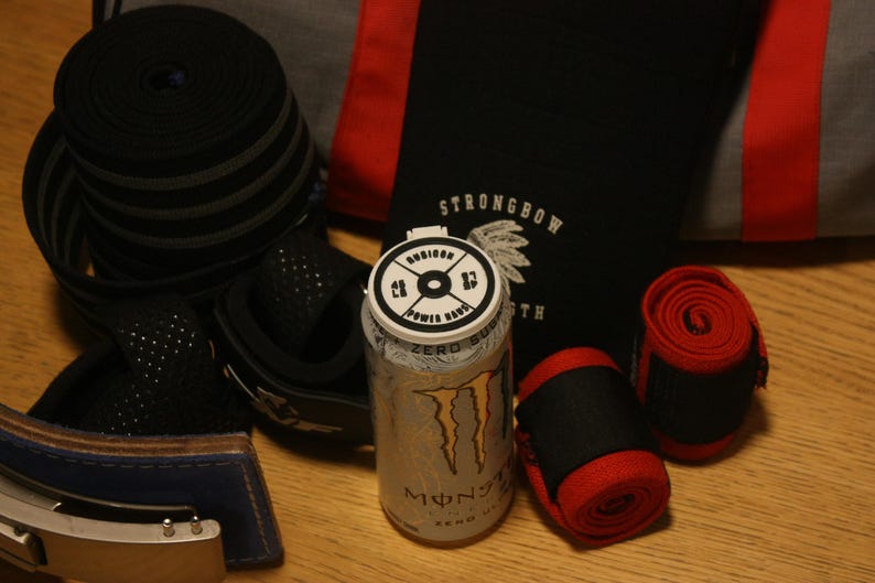 May include: An assortment of fitness equipment, including black and red wrist wraps, a weightlifting belt, and a rolled-up black band. A silver Monster Energy drink can is also present, along with a black bag with the text "STRONGBOW".