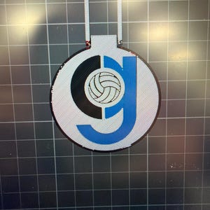May include: A silver ornament with a volleyball design. The ornament features a black and blue letter "G" with a volleyball in the center. The ornament has a loop at the top for hanging.