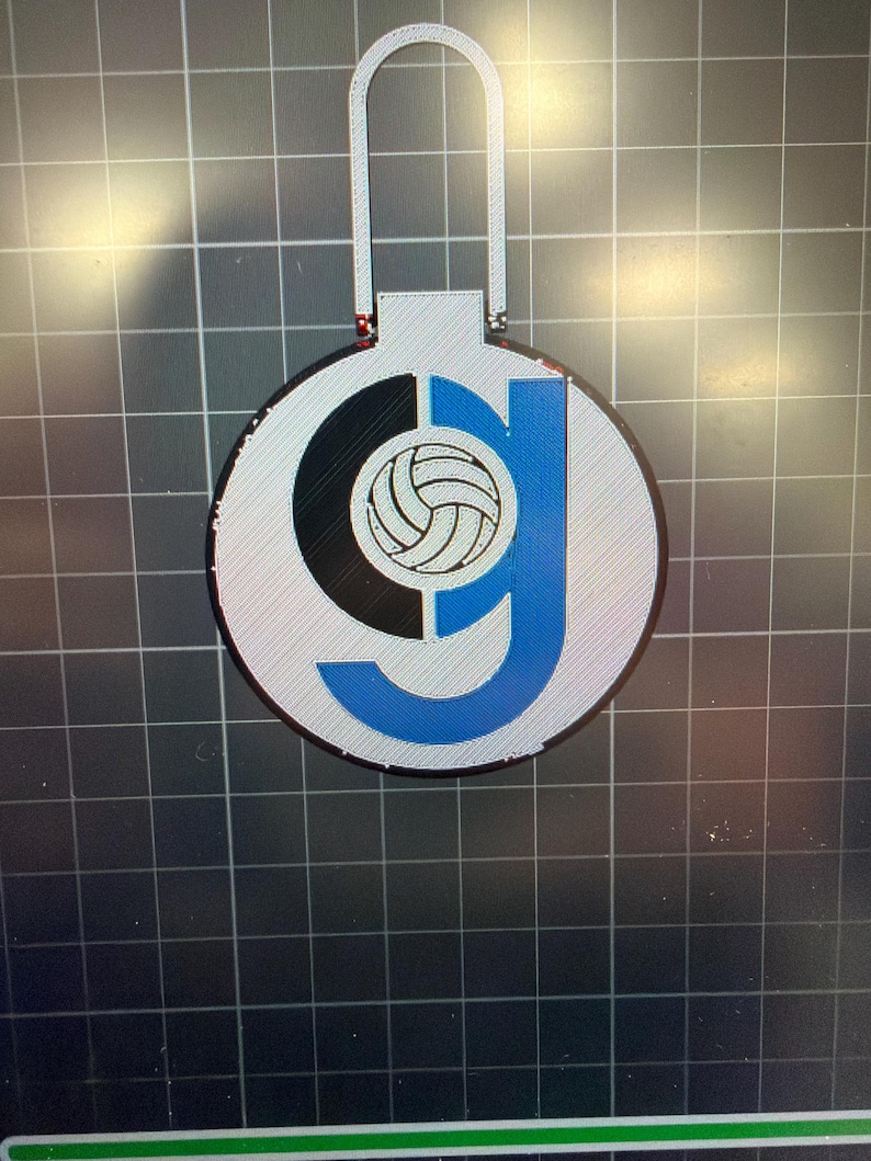 May include: A silver ornament with a volleyball design. The ornament features a black and blue letter "G" with a volleyball in the center. The ornament has a loop at the top for hanging.