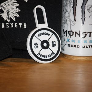 May include: A white and black weight plate keychain with the text "RUBICON" and "POWER HAUS". The keychain has a loop at the top. A black bag with the word "STRENGTH" and a Native American headdress graphic is in the background.