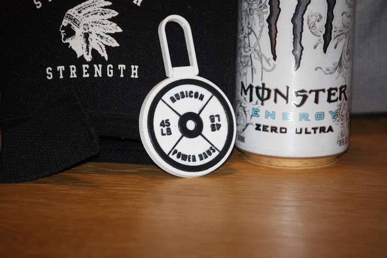 May include: A white and black weight plate keychain with the text "RUBICON" and "POWER HAUS". The keychain has a loop at the top. A black bag with the word "STRENGTH" and a Native American headdress graphic is in the background.