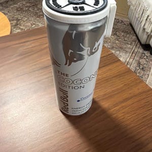 May include: A silver Red Bull can with a white lid and handle, featuring the text "THE COCONUT EDITION." The can has a silver bull logo and additional text indicating the drink's contents. The can is on a wooden surface.