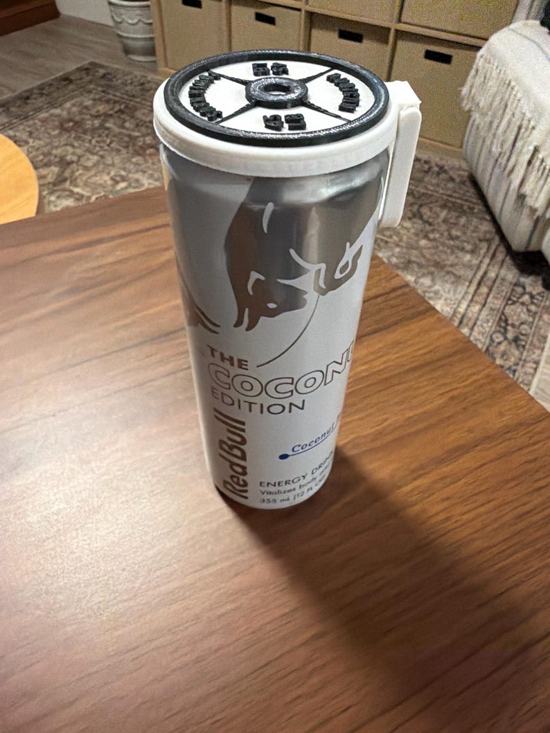 May include: A silver Red Bull can with a white lid and handle, featuring the text "THE COCONUT EDITION." The can has a silver bull logo and additional text indicating the drink's contents. The can is on a wooden surface.