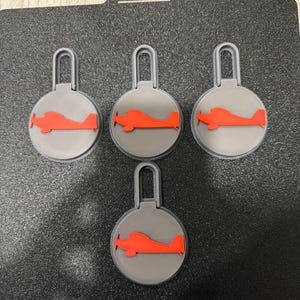 May include: Four gray keychains with a red airplane design. Each keychain has a circular base with a red airplane silhouette and a gray loop at the top. The keychains are arranged on a black surface.