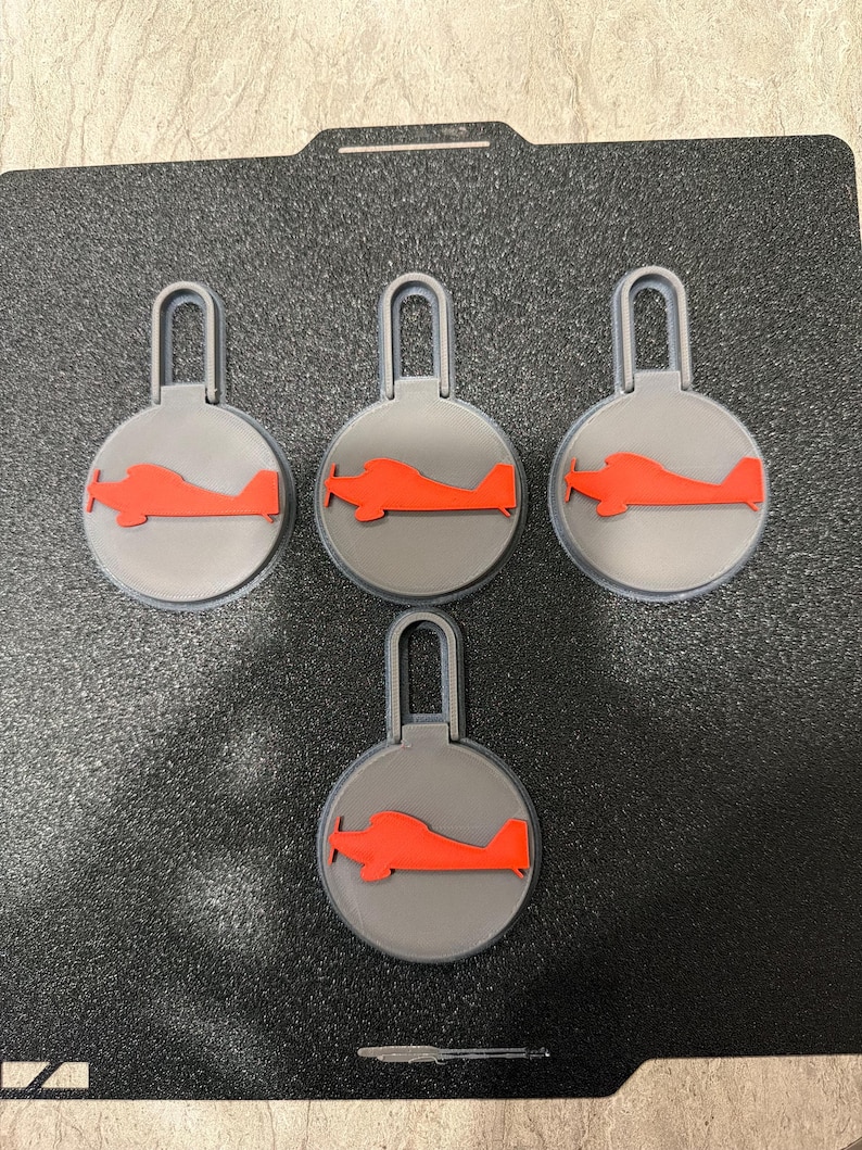 May include: Four gray keychains with a red airplane design. Each keychain has a circular base with a red airplane silhouette and a gray loop at the top. The keychains are arranged on a black surface.