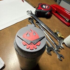 May include: A gray can with a red and black flame design on the lid. The can is surrounded by tools, including wrenches and a tape measure. The can has the word "MONSTER" printed on it.