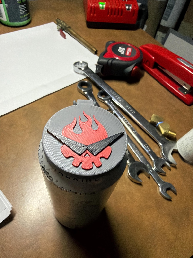May include: A gray can with a red and black flame design on the lid. The can is surrounded by tools, including wrenches and a tape measure. The can has the word "MONSTER" printed on it.