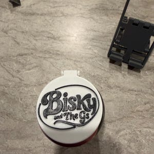 May include: A white and black round object with the text "Biskys and The Gs" in a black, stylized font. The object has a hinged lid and a red base. A utility knife and a black plastic frame are in the background.