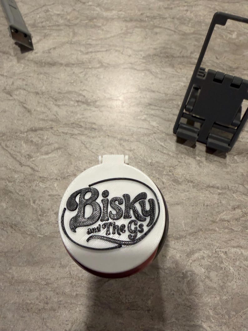 May include: A white and black round object with the text "Biskys and The Gs" in a black, stylized font. The object has a hinged lid and a red base. A utility knife and a black plastic frame are in the background.