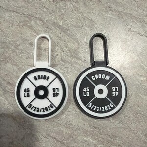 May include: Two weight plate-shaped wedding favors. One is white with "BRIDE" and the other is black with "GROOM". Each has "45 LB" and "87 SP" with the date "5/23/2026".