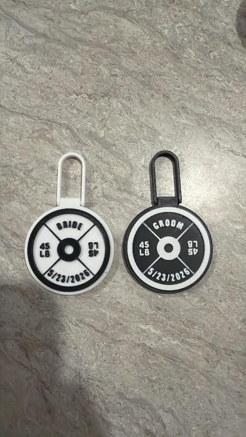 May include: Two weight plate-shaped wedding favors. One is white with "BRIDE" and the other is black with "GROOM". Each has "45 LB" and "87 SP" with the date "5/23/2026".