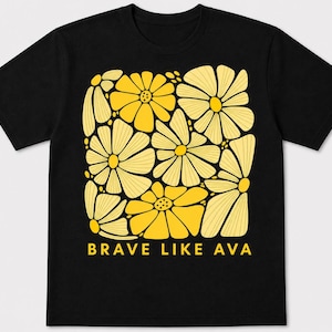 May include: Black t-shirt featuring a square design of yellow flowers with a light yellow outline. The text "BRAVE LIKE AVA" is printed in yellow below the floral design. The shirt is made of a soft material.