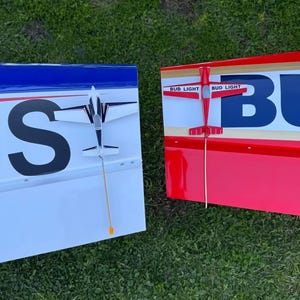 May include: Two model airplanes, one white and blue, the other red and white, rest on grass. The white and blue plane has the letters "NS" on its wing. The red and white plane has "BUD LIGHT" printed on its wing.