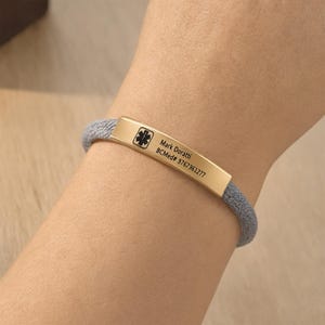 May include: A medical alert bracelet featuring a gold-toned metal bar with a black Star of Life symbol, the name Mark Doratti, and the text BCMed# 9767361277 engraved on the surface, attached to a textured grey cord band.