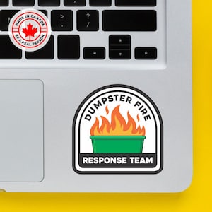 Peut inclure: Un autocollant avec le texte "DUMPSTER FIRE RESPONSE TEAM" et une illustration d'une poubelle verte avec des flammes orange. L'autocollant est sur un clavier d'ordinateur portable. Un petit autocollant indique "MADE IN CANADA BY A REAL PERSON."