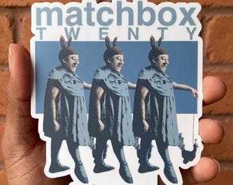May include: A die-cut sticker featuring the text matchbox TWENTY at the top and mad season at the bottom. The central graphic displays three identical figures in blue tones, each wearing a hooded cloak with rabbit ears, with one figure holding a small bird.